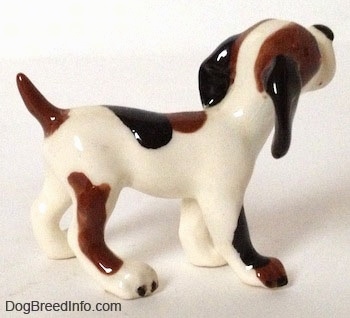 The right side of a vintage white with brown and black Hound dog figurine. The figurine has  medium-sized legs and big paws. The body is mostly white and the ears are black. There are black and brown patches on it. The tail is brown.