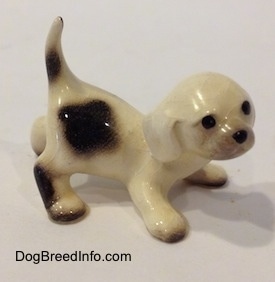 The right side of a white with brown Hound puppy figurine that is crouching. The figurine has black circles for eyes and a nose.