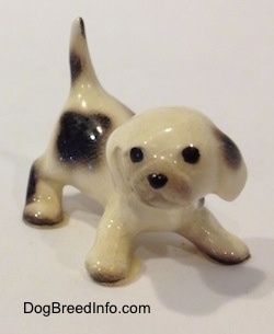 The front right side of a white with brown spotted Hound puppy figurine in a crouching position. The figurine has its tail in the air.