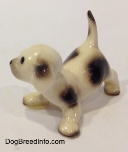 The front left side of a brown spotted Hound puppy in a crouching position figurine. The figurine has a black spot on its white ear.