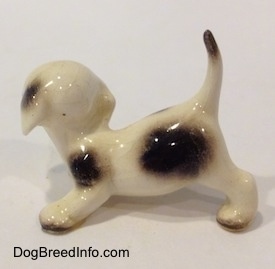 The left side of a white with brown spotted Hound puppy crouching figurine. The figurine has two brown spots one near its hind legs and one above its front legs.