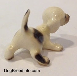 The back right side of a white crouching hound puppy figurine that has brown spots.