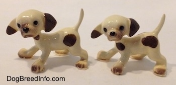 The left side of two white with brown spotted Hound puppy figurines. Both figurines has black circles for eyes.