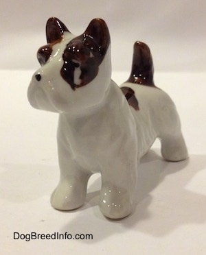 The front left side of a white with brown bone china Jack Russell Terrier dog figurine. The figurine has a large chip under its eye.