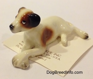 The front left side of a figurine of a white with brown and black Jack Russell Terrier dog in a lying down pose. The eyes of the figurine are very cartoon-y