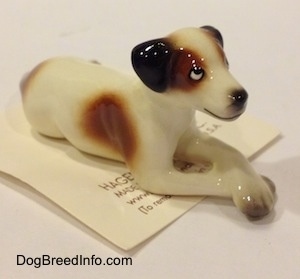 The front right side of a white with brown and black Jack Russell Terrier dog in a lying down pose figurine. The figurine has a black line for a mouth.