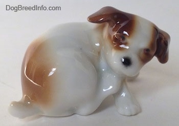The right side of a Jack Russell Terrier figurine that is scratching its neck. The figurine has small black circles for eyes.