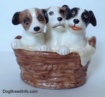 A figurine of three Jack Russell Terriers that are in a wicker basket. The figurine on the right has its mouth open.