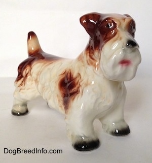 The front right side of a white with brown Lucas Terrier figurine. The figurine has a pink mouth and black circles for eyes.