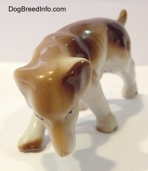 A brown and whtie Lurcher figurine is sniffing the ground and it has black circles for eyes.