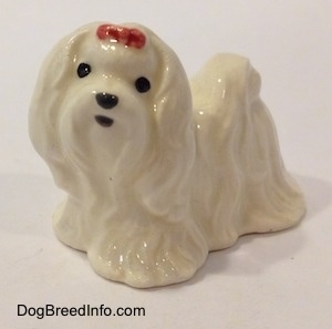 The front left side of a white Maltese figurine with a red ribbon in its hair. The figurine has black circles for eyes and a nose.