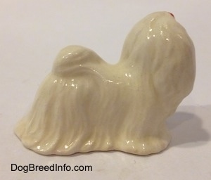 The right side of a white Maltese figurine with a red ribbon in its hair. The tail of the figurine has its tail curled on its back.