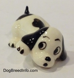 The front right side of a figurine of a white with black puppy figurine that is in a lying pose. The figurine has black circles for eyes and a nose.
