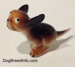A figurine of a miniature running puppy that is brown and black with white. The figurine has black circles for eyes, it is looking up and to the right.