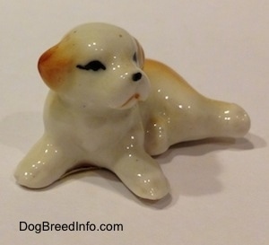 The front left side of a white with brown bone china puppy figurine that is in a lying down pose. The figurine has short legs.
