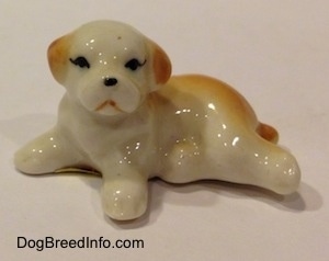 A white with brown bone china puppy figurine that is in a lying down pose. The figurine has black circles for eyes.