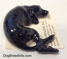 A figurine of a black dog with grey highlights that is in a sleeping pose. The figurine has a long tail.