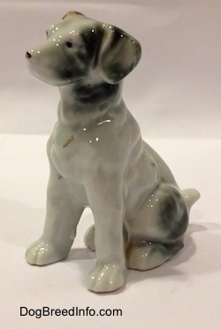 The front left side of a bone china brown, white and black figurine of a mixed breed dog in a sitting pose. The figurine has long limbs.