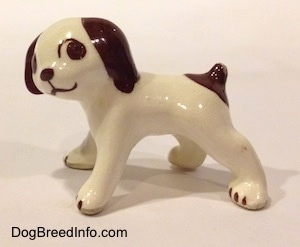 The left side of a white with brown dog standing figurine. The figurine has a short brown tail with a brown spot around it.