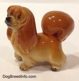 The left side of a figurine of a brown with tan Pekingese puppy. The ears of the figurine are hard to differentiate from its head and body.