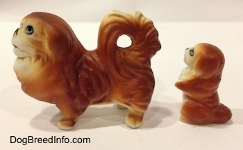 The left side of a brown with white Pekingese figurine and behind it is a brown with white Pekingese puppy in a begging pose figurine. The figurines have painted faces.
