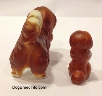 The back of a figurine of a Pekingese puppy in a begging pose and a figurine of a brown with white Pekingese figurine. The tails of the figurines are arched up on each others back.