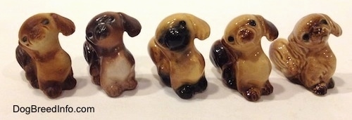 The front right side of five different color variations of a figurine of a Pekingese puppy seated figurine. The figurines have black circles for eyes.
