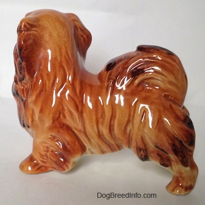 The left side of a figurine of a brown and tan with black Pekingese. The figurine has its tail curled onto its back.