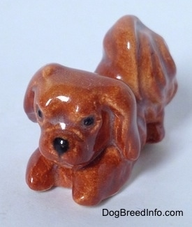 The front left side of a figurine of a brown Pekingese that is in a play bow pose. The figurine has black circles for eyes.