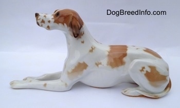 The left side of a porcelain figurine of a white with brown Pointer in a lying pose. The figurine has long legs.