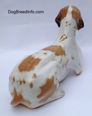 The back right side of a porcelain white with brown figurine of a Pointer in a lying pose. The figurine has a long tail along its left side.