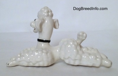 The left side of a white figurine of a Poodle in a lying position. The figurine has a short tail with a large hair poof at the end.