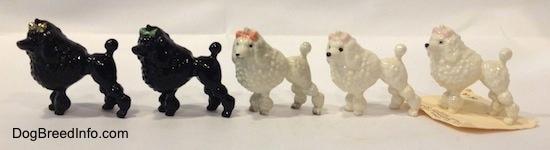 The left side of five different color variations of a figurine of a Poodle wth a bow in its hair. The figurines have black circles for eyes.