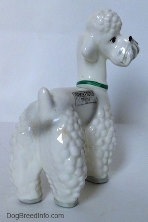 The back right side of a figurine of a white Poodle with a green collar on. The hair around the figurines mouth makes it look like it has a Moustache.