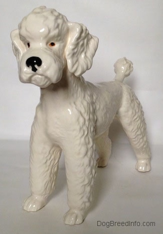 The front left side of a figurine of a white Poodle. The eyes of the figurine are black and orange.