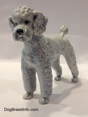 The front left side of a figurine of a porcelain white with blue Poodle standing. The figurine has black circles for eyes and a glossy nose.