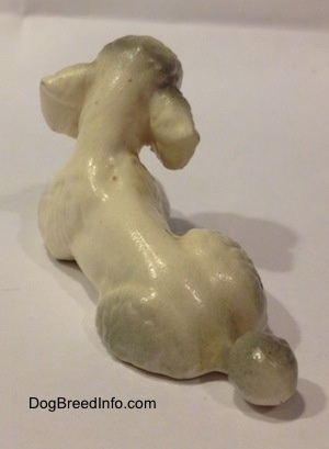 The back left side of a white figurine of a ceramic Poodle in a lying position. The figurine has a small tail with a large black tinted poof at the end.