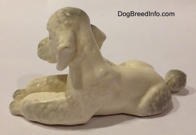 The left side of a ceramic white Poodle figurine in a lying position. The figurine has a poof of hair on its head.
