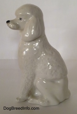 The left side of a porcelain white standard Poodle figurine in a sitting pose. The figurine has very fine hair details along its body and front legs.