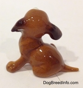 The left side of a brown with white figurine of a miniature puppy sitting. The figurine has a short tail.