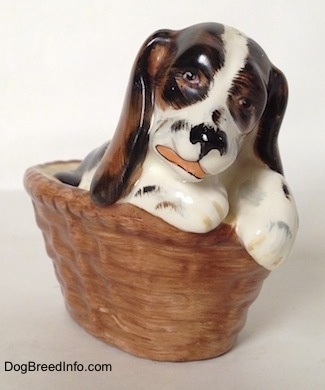 A white with brown and black figurine of a Russian Spaniel puppy in a basket. The figurine has detailed eyes.