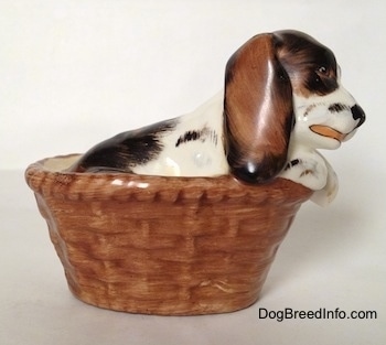 The right side of a white with brown and black figurine in a basket of a Russian Spaniel puppy. The figurine has brown ears with black edges.