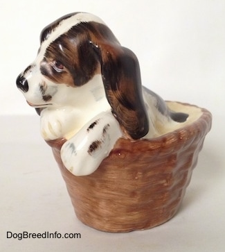 The front left side of a white with brown and black Russian Spaniel puppy in a basket figurine. The figurine has its paws on the edge of the basket.