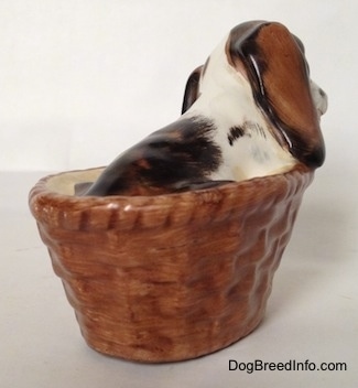 The back right side of a white with brown and black Russian Spaniel puppy in a basket figurine. The figurine has its head tilted towards the left.
