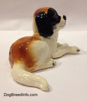 The back right side of a white and brown with black Saint Bernard puppy figurine in a laying down position. The figurine has a large brown spot across its back and towards its tail.