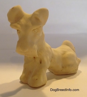 The front left side of a figurine of a Miniature Schnauzer sitting. The figurine is made out of clay.