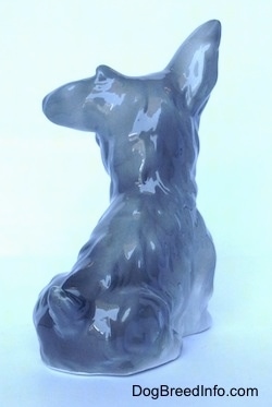 The back right side of a figurine of a grey and white figurine of a miniature Schnauzer sitting. The figurine is glossy.