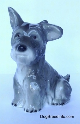 The front left side of a grey and white figurine of a miniature Schnauzer sitting. The figurines left ear is hanging to the side and its right ear is in the air.