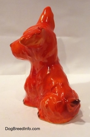 The back left side of a figurine of an orange miniature Schnauzer sitting. The tail of figurine is arched up and it has a black tip.