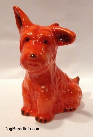 The front left side of an orange miniature Schnauzer figurine that is in a sitting pose. The figurine has black circles for eyes and a nose.